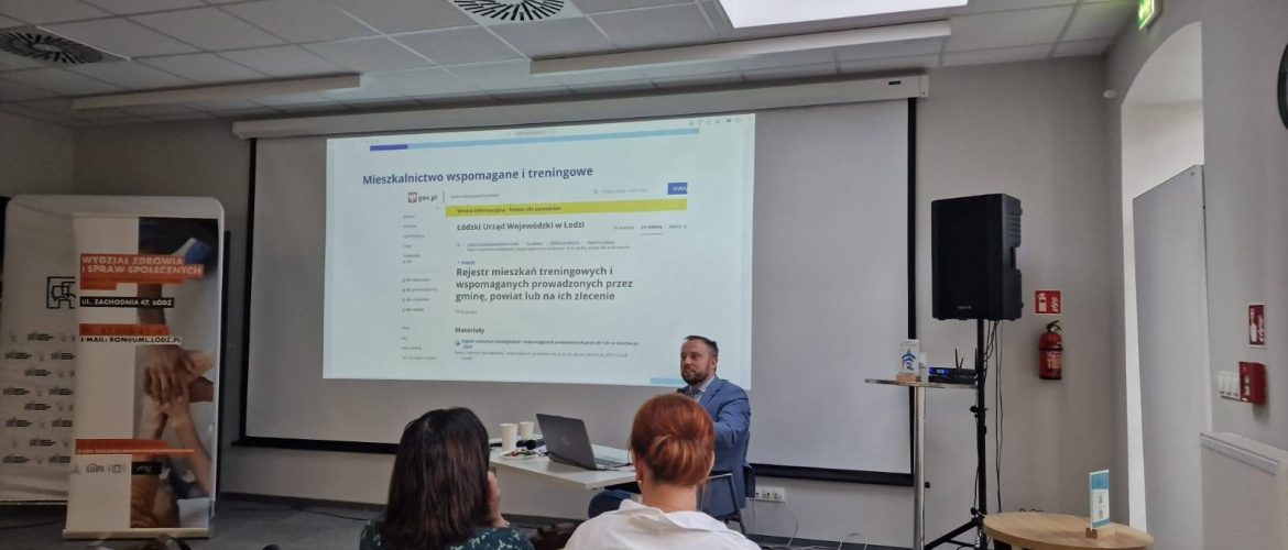 Na zdjęciu widzimy mężczyznę ubranego w granatowy garnitur, siedzącego przy biurku. Na tablicy multimedialnej wyświetlane są treści warsztatów.