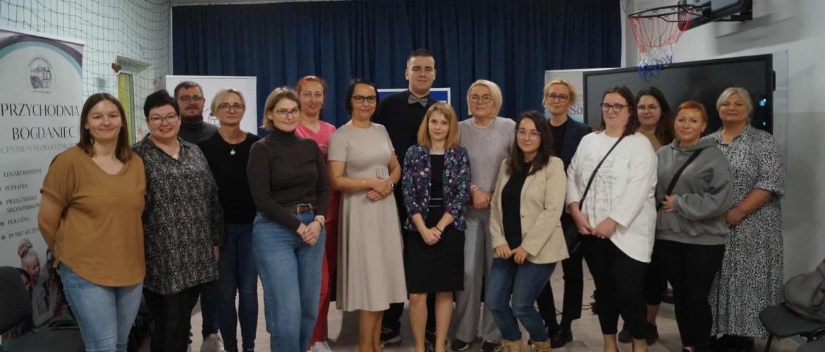 Na zdjęciu widzimy uczestniczki oraz prowadzących spotkanie stojących przy tablicy imultimedialnej oraz banerze, na którym jest napisane "Przychodnia Bogdaniec".