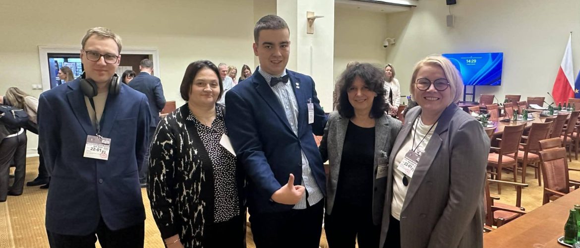 Grupa pięciu osób z organizacji społecznych stoi w sali konferencyjnej im. Jacka Kuronia, podczas posiedzenia Podkomisji Stałej ds. Zabezpieczenia Społecznego. W tle widoczne są stoły konferencyjne, krzesła oraz flaga Polski.