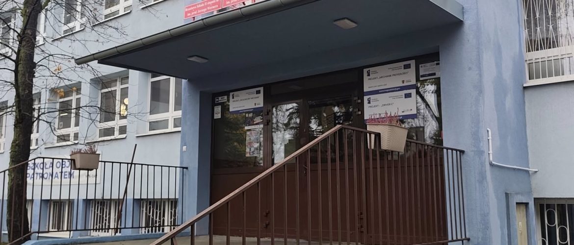 „Wejście do Zespołu Szkół Samochodowych w Łodzi. Nad drzwiami znajduje się czerwony Fiat 126p umieszczony na daszku. Budynek jest niebieski, z kilkoma oknami i schodami prowadzącymi do wejścia.”