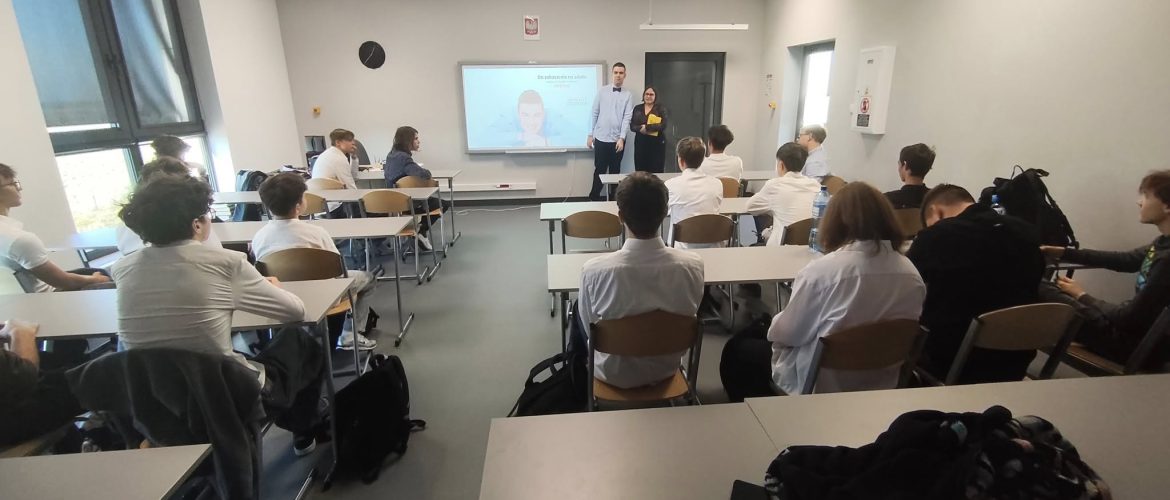 „Sala lekcyjna w nowoczesnej szkole, w której odbywa się spotkanie w ramach Lekcji Różnorodności. Uczniowie siedzą przy stołach, zwróceni w stronę ekranu z prezentacją. Na ekranie widoczny jest slajd z napisem „Neurodiversity Matters”. Przed ekranem stoją dwie osoby prowadzące zajęcia, rozmawiające z grupą.”