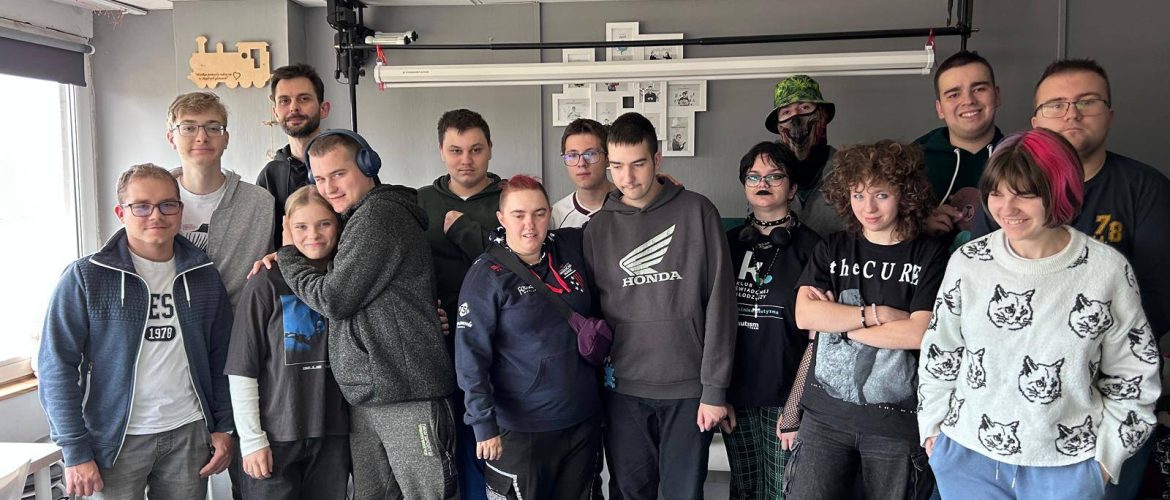 Grupa samorzeczniczek i samorzeczników stoi w pomieszczeniu o jasnych ścianach i podwieszanym suficie z panelami. W tle znajduje się tablica lub ekran zamocowany na ścianie, nad którym widoczne są kartki z rysunkami lub schematami. Na pierwszym planie stoi kilka białych plastikowych krzeseł oraz stół z rozłożonymi kartkami, rysunkami i przyborami do pisania, w tym zielonym folderem. Pomieszczenie jest dobrze oświetlone światłem dziennym wpadającym przez okno po lewej stronie.